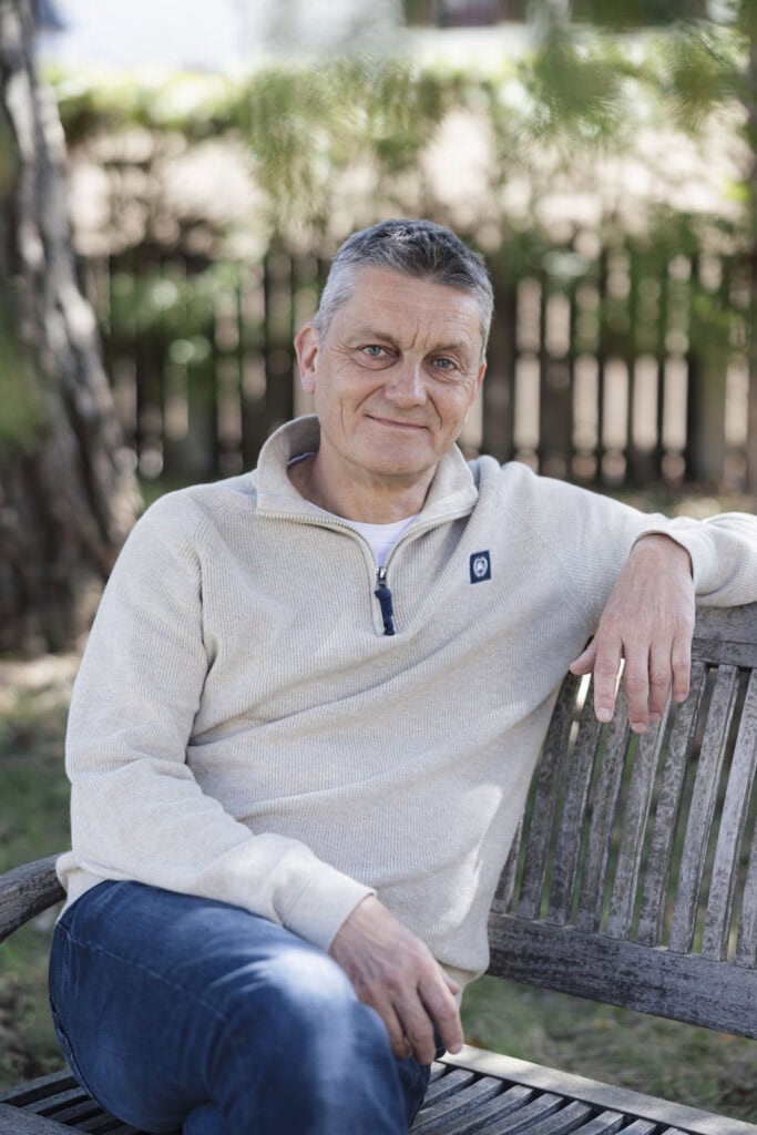 Gerhard Riedle entspannt auf einer Parkbank sitzend, Natur im Hintergrund.